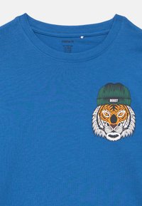 Μπλε μπλούζα με στρογγυλή λαιμόκοψη, με εκτυπωμένο πρόσωπο τίγρης που φοράει πράσινο σκουφάκι με την ετικέτα "BEAST" στο πάνω αριστερό μέρος του στήθους.