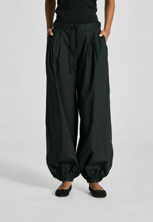 Personne portant un pantalon noir ample avec cordon de serrage et poignets élastiques froncés, ainsi que des ballerines noires, debout les mains dans les poches.