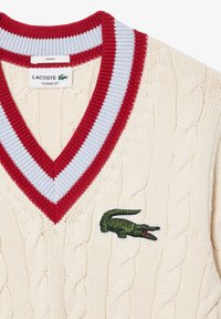 Pullover de malha em creme com decote em V, apresentando riscas em vermelho e azul claro nas margens e um logótipo de crocodilo bordado em verde no peito.