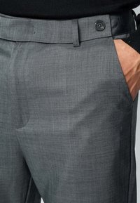 Pantalon gris sur mesure en tissu lisse, avec une fermeture à boutons, des poches latérales et un motif texturé subtil.