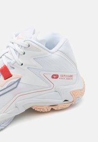 Adidași albi cu un superior texturat, accente roșii și piersici moi, și un design ondulat distinctiv pe talpă pentru o aderență suplimentară.