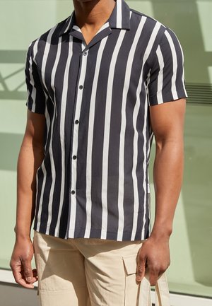Homme portant une chemise à manches courtes rayée verticalement noir et blanc et un short cargo beige tenant un sac fourre-tout beige.