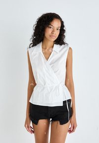 White sleeveless wrap top with a collared neckline, cinched waist, and peplum design, paired with black distressed denim shorts.