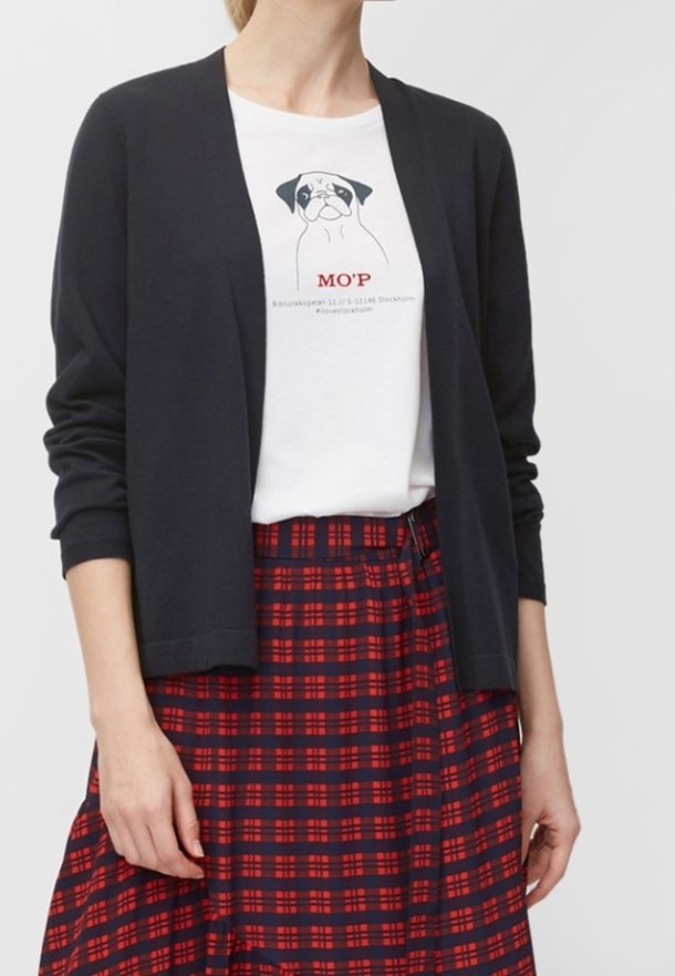 Gilet noir à ouverture frontale et à manches longues, porté sur un t-shirt graphique blanc et une jupe écossaise rouge et marine. Texture lisse et coupe décontractée.
