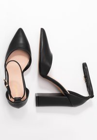 Tacchi slingback in pelle nera con punta affusolata, tacco a blocco spesso e cinturino alla caviglia con fibbia in tonalità oro. Texture liscia e design minimalista.