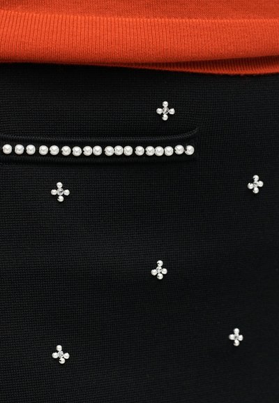 Μαύρο ύφασμα με μικρές συσσωρευμένες πέρλες και στρας, μια οριζόντια σειρά από πέρλες πάνω από μια τσέπη και μια κόκκινη πλεκτή άκρη ενδύματος από πάνω.