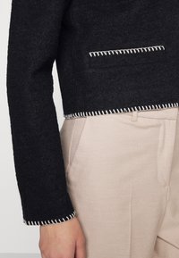 Czarna krótka kurtka z teksturowanej tkaniny, z białymi stębnowaniami na dolnym brzegu, zestawiona z jasnobeżowymi spodniami w kant.