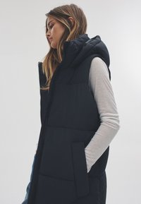 Fekete puffertöltéses mellény kapucnival, cipzáros elülső résszel, oldalzsebekkel és steppelt mintázattal. Egy világosszürke hosszú ujjú póló fölött viselve.