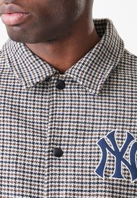 Κοντινή λήψη ενός ατόμου που φοράει ένα μπουφάν με καρό σχέδιο (houndstooth) με μαύρα κουμπιά και ένα μπλε και λευκό κεντητό λογότυπο των New York Yankees.