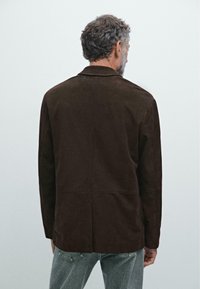Giacca in suede marrone con colletto a punte, design senza cuciture e tasche laterali, mostrata dal retro su uno sfondo chiaro.