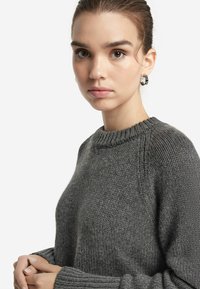 Grauer Pullover mit einem gerippten Rundhalsausschnitt, strukturiertem Strickmuster und lockeren Ärmeln. Das Material wirkt weich und leicht dick.