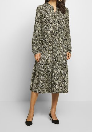 Robe noire avec un motif floral blanc et jaune, manches longues, col boutonné et une jupe à volants, associée à des talons noirs.