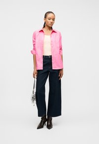 Γυναίκα με πλεξούδες, που φοράει ριγέ ροζ πουκάμισο, κρεμ τοπ, σκουρόχρωμο τζιν παντελόνι φαρδιάς γραμμής, μαύρες μπότες και κρατάει ασημένια τσάντα, μπροστά σε λευκό φόντο.