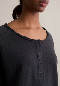 Ciemnoszara koszulka henley z postarzanym okrągłym dekoltem, sześcioma guzikami i długimi rękawami, wykonana z miękkiej, teksturowanej tkaniny.
