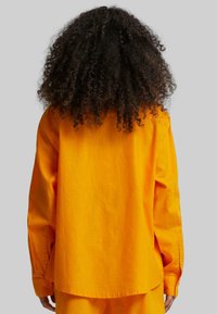 Långärmad skjorta i klar orange, gjord av strukturerad tyg, med avslappnad passform, knäppningar vid ärmsluten och rundad nederkant. Bakvy.