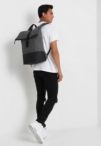 Zaino in tessuto grigio con base nera, spallacci regolabili e chiusura a rotolo. Il materiale sembra tessurato con un design minimalista.