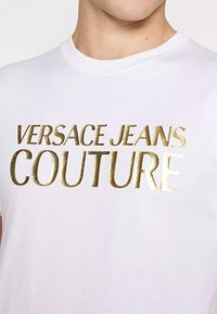 T-shirt bianca con stampa metallica dorata "VERSACE JEANS COUTURE." Design classico a girocollo con maniche corte e texture liscia.