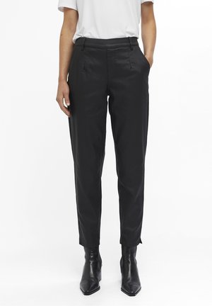 Personne portant un pantalon noir sur mesure longueur cheville, des bottes noires à bout pointu et une chemise blanche à manches courtes, debout les mains dans les poches.