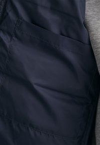Gros plan d'une poche en tissu bleu marine foncé sur un vêtement avec des coutures visibles et une partie d'une manche grise à côté.