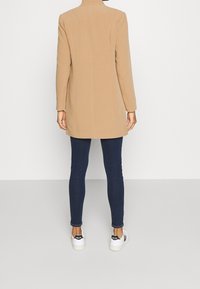 Tan, mellanhängig kappa med hög krage och skräddarsydd passform, i kombination med mörkblå slim fit jeans och vita sneakers med svarta detaljer.