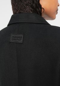 Casaco de lã preto com colarinho estruturado e acabamento texturizado. Inclui um emblema da marca "Fabienne Chapot" nas costas.