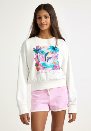 Teenagepige iført hvid sweatshirt med tropisk delfin- og palmeprint med teksten "Hidden Lagoon" og lyserøde shorts.
