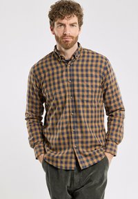 Homme avec des cheveux bouclés et une barbe, portant une chemise à carreaux marron et marine à boutons et un pantalon en velours côtelé vert foncé, les mains dans les poches.