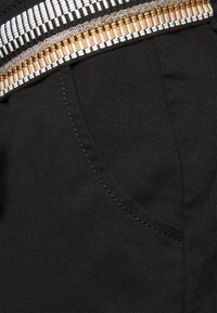 Tissu noir à surface texturée, avec une ceinture tissée détaillée en tons or, blanc et noir. Poche latérale visible.