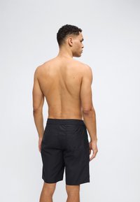 Άνδρας χωρίς πουκάμισο με κοντά, σγουρά μαλλιά, φορώντας μαύρο μαγιό, στεκόμενος πλάτη σε απλό ανοιχτό γκρι φόντο.