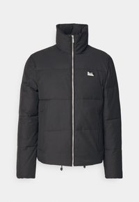 Sort pufjakke med høj krave, frontlynlås, quiltet design, elastisk kant og et lille logo-mærke på brystet.