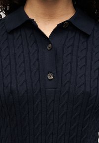 Πλεκτό μπλε κοντομάνικο πόλο πουκάμισο με ανάγλυφο σχέδιο καλωδίου, γιακά και τρία σκούρα κουμπιά στον λαιμό, με κοντινή άποψη του υφάσματος.