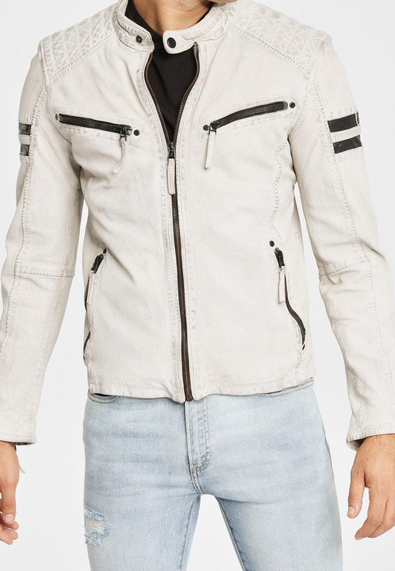 Homme portant une veste blanche à fermeture éclair avec des poches zippées noires et des détails en bandes sur les manches, associée à un jean bleu clair déchiré.
