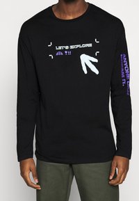 Svart långärmad tröja med grafisk text "Låt oss utforska" och symboler. Accenter i ljusblått och lila på ärmarna. Material i bomull.