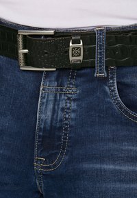 Schwarzer Leder-Gürtel mit Krokodilmuster, silberner Schnalle und Beschlägen, getragen über dunkelblauen Jeans mit kontrastierenden Nähten.