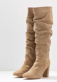 Botas de ante en color beige hasta la rodilla, holgadas, con punta afilada y tacón en bloque. La textura es suave, con costuras visibles a lo largo del caño.