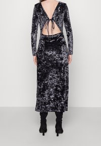Robe longue en velours noir à manches longues avec une découpe profonde dans le dos, nouée à la taille. Texture lisse avec un léger éclat et silhouette ajustée.