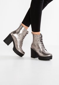 Metallic sølv ankelstøvler med en kraftig sort platform og hæl, der har snørebånd og en pull tab, præsenteret mod en hvid baggrund.