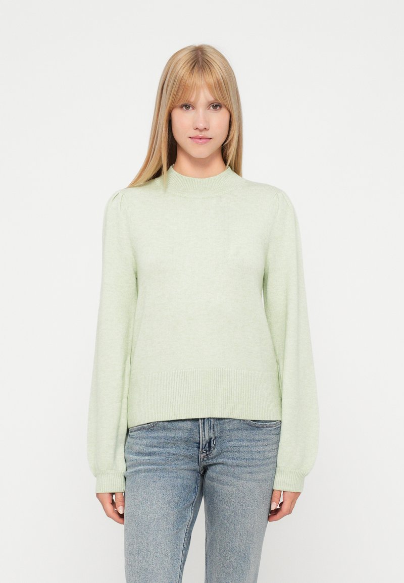 Gaiši zaļa adīta džemperis ar augstu kakla izgriezumu,  puffām piedurknēm un ribotu piedurkņu un apakšmalu. Apvienots ar ziliem džinsiem.