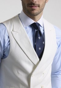 Chaleco blanco de doble botonadura sobre una camisa azul clara, complementado con una corbata azul oscura con un sutil patrón de heráldica. Textura de tela suave.