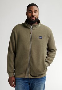 Olivgrön zip-up jacka med ett strukturerat mönster, hög krage och sidofickor. Har en liten logotyp-patch på bröstet.