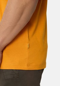 Oranges Baumwoll-T-Shirt mit seitlichem Nahtetikett, kurzen Ärmeln und geradem Saum. Die Stoffstruktur wirkt glatt.