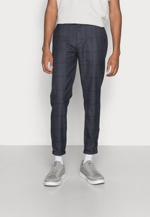 Uomo che indossa pantaloni skinny a quadri grigio scuro, calzini bianchi, scarpe da ginnastica grigie e una camicia bianca a maniche corte, in piedi davanti a uno sfondo neutro.