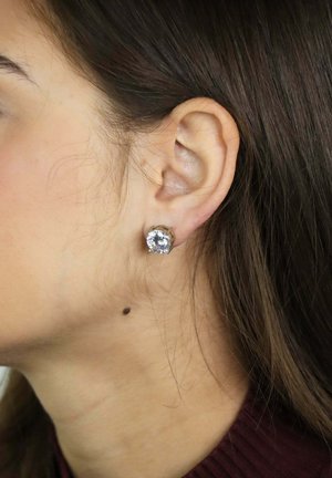 Linkeroor van een vrouw met een grote ronde oorbel met een heldere edelsteen en een klein donker moedervlekje op haar nek onder het oor.