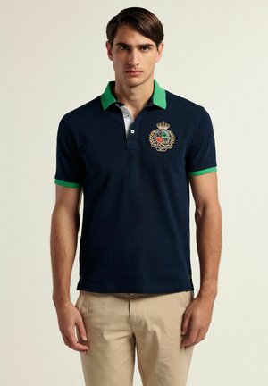 Polo de color marino con cuello verde y blanco, emblema bordado, mangas cortas y detalles a rayas en el dobladillo. Material de algodón.