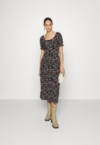 Vestido preto com padrão floral, mangas bufantes, cintura ajustada e saia até ao joelho; combinado com botas de salto bloco bege e uma clutch creme.