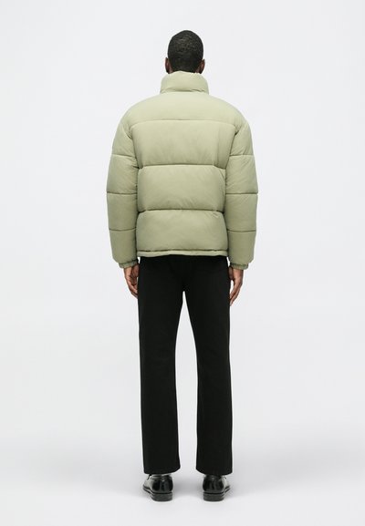 Kurtka puchowa w jasnym oliwkowym zielonym kolorze, z wysokim kołnierzem i pikowanym wzorem. Czarne spodnie i buty dopełniają stylizację.