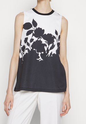 Vrouw draagt een mouwloze zwart-witte top met zwarte bloemenprint op de bovenste helft en een witte broek tegen een lichte achtergrond.