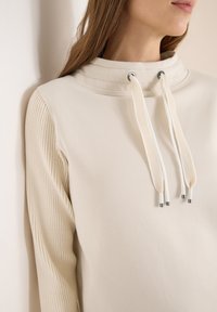 Felpa color crema con colletto alto, coulisse con dettagli in argento e maniche a coste per una consistenza maggiore.