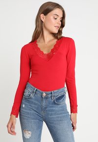 Body rosso a maniche lunghe con scollo a V e bordi in pizzo, design aderente e dettagli in pizzo sui polsini, abbinato a jeans strappati di un blu chiaro.
