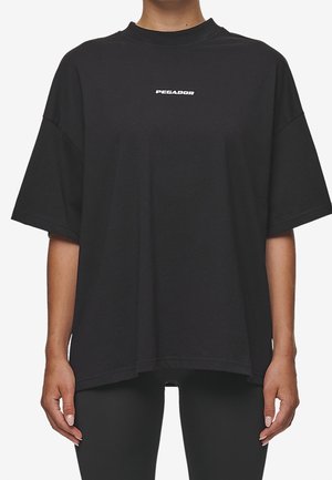 Persoon draagt een oversized zwart T-shirt met korte mouwen en witte tekst "PEGADOR" gecentreerd op de borst.